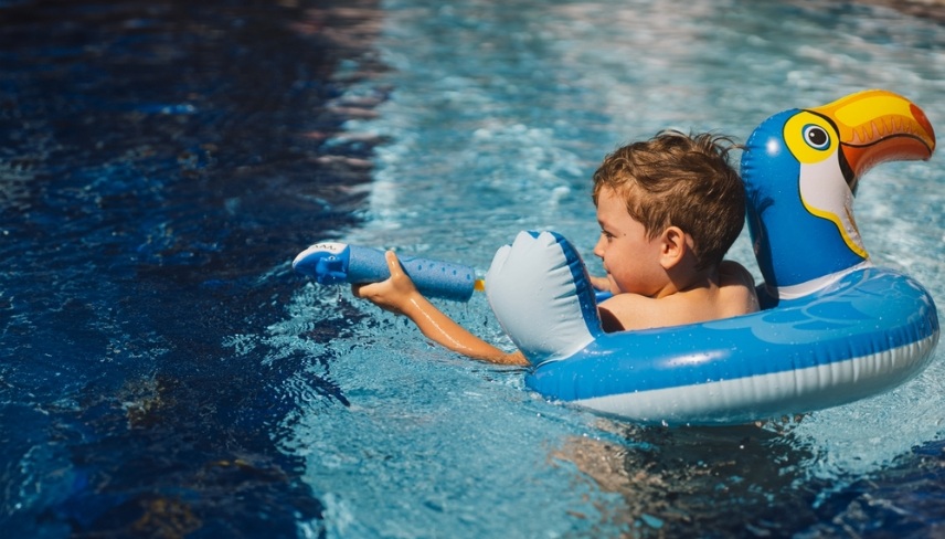 A boy with a joyful expression is floating in a swimming pool on a sunny day, using an swimming circle designed to look like a blue bird.