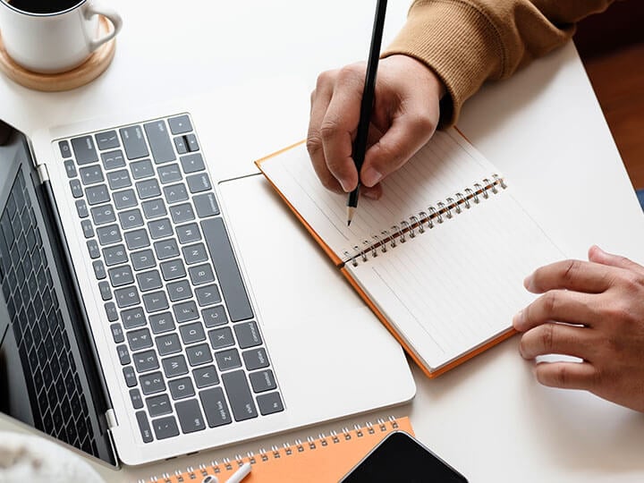 Aerial view of adult male writing in a notebook with his laptop out on a desk and a cup of coffee next to it